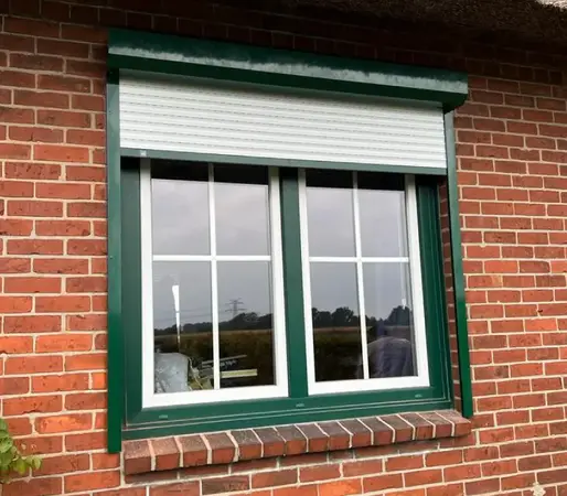 Grünes Kunststofffenster mit Rollladen in roter Klinkerfassade eines Hauses.